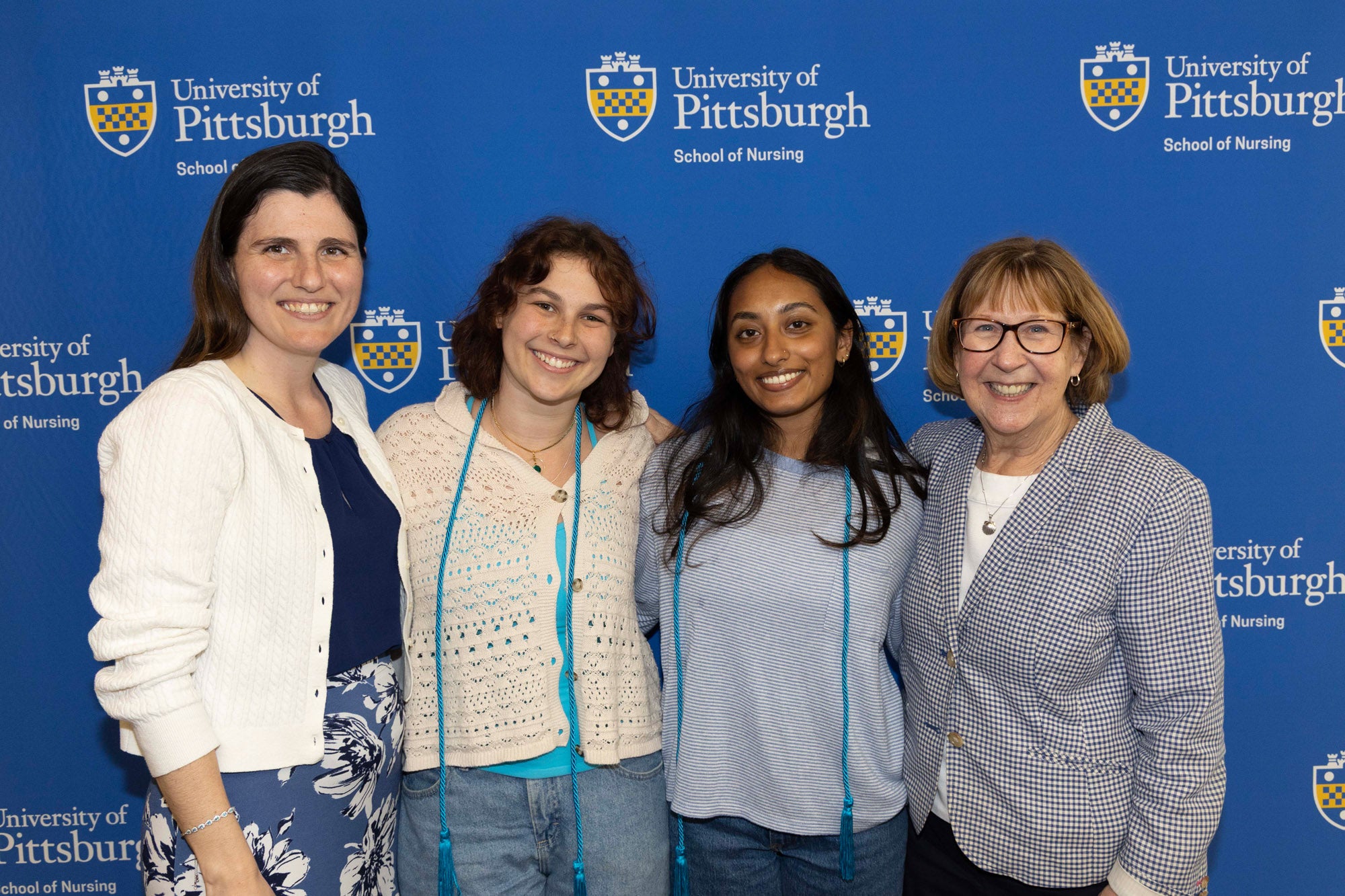 Group of four women smile at the camera as they stand in front of a blue background emblazoned with the University of Pittsburgh logo