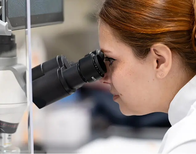 Nurse with hair net and surgical mask prepares to view a specimen in a microscope.