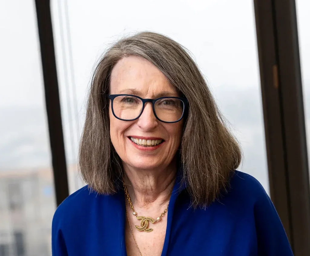 Photo of Christine Kasper, smiling in a blue dress, wearing glasses sitting in front of a window
