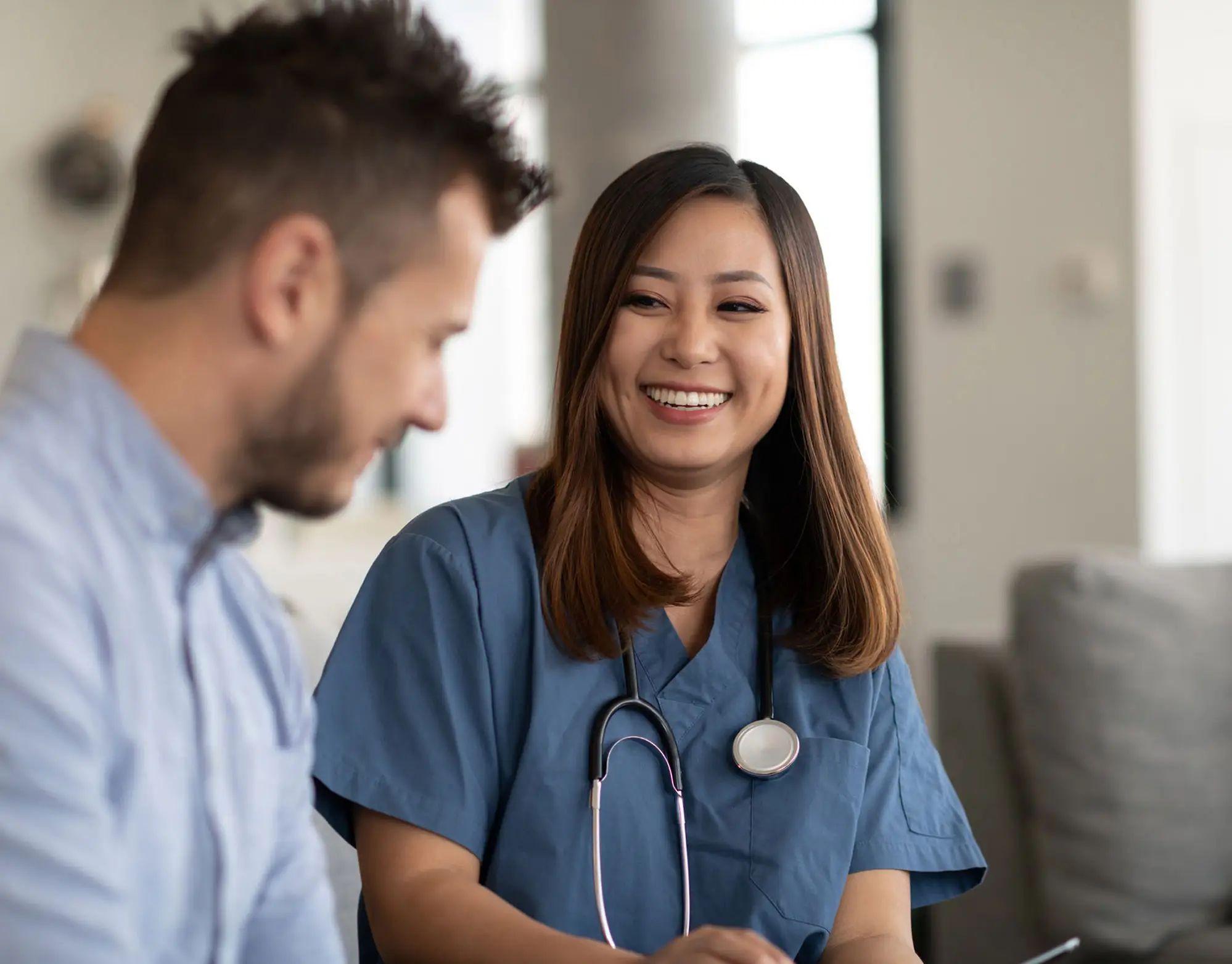 A young doctor of Asian descent and a young Caucasian patient of are indoors at the man's home. The doctor is talking to the man while holding a tablet computer.